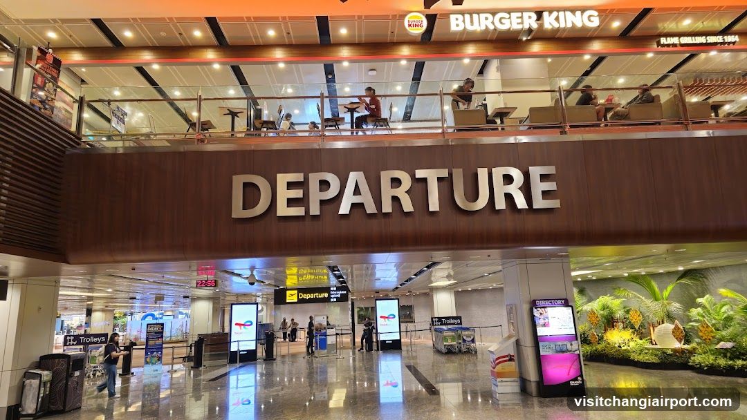 Interior view of an airport departure hall with a large 'DEPARTURE' sign above the ground floor. Upper level features Burger King and Flame Grilling restaurants with customers dining at tables. Ground floor includes baggage claim trolleys, information screens, and a landscaped garden area.
