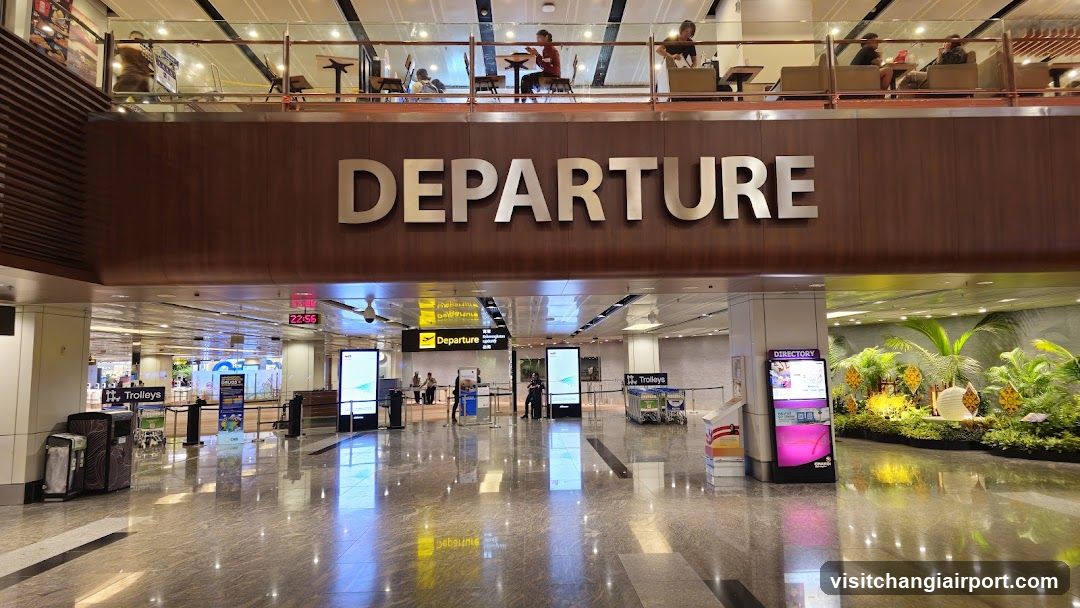 Interior view of an airport departure hall with polished marble floors, a large 'DEPARTURE' sign above the gate area, and a mezzanine level with seating. Visible elements include trolley bins, digital screens, directional signs, and tropical plant displays.