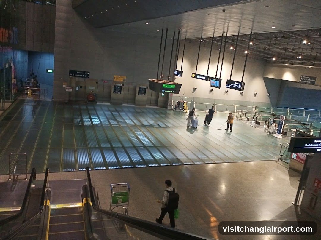 A spacious airport terminal with polished floors, hanging digital screens displaying flight information, and a few travelers navigating the area. The scene includes an escalator in the foreground and signage indicating directions to terminals.