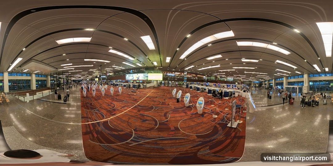 A panoramic view of a spacious airport terminal with high ceilings, curved architectural elements, and rows of self-service kiosks on patterned carpeting. Passengers are visible in the background near check-in counters and seating areas.