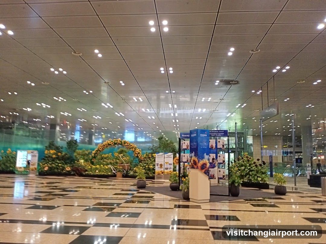A spacious indoor area with a polished tiled floor and a high ceiling with recessed lighting. The space features decorative elements including sunflower arrangements, greenery, and informational posters. There are no visible shop fronts or menus.