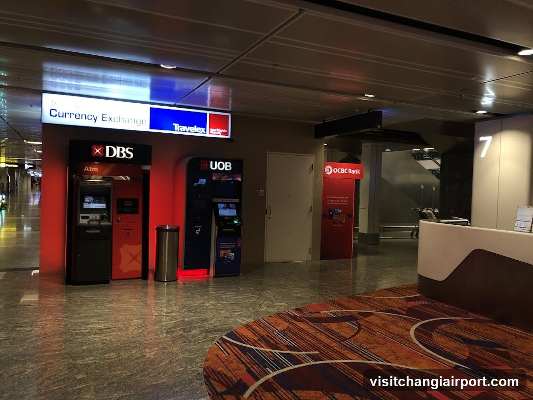 Interior view of an airport terminal showing currency exchange kiosks (DBS, UOB) with red lighting accents, a trash bin, and signage for OCBC Bank in the background. A patterned carpet is visible in the foreground.