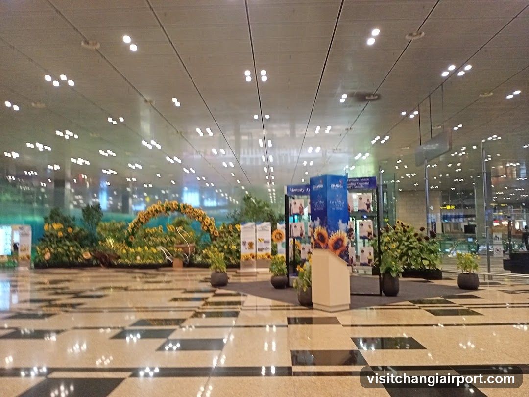 A spacious airport terminal hall featuring polished checkered flooring and a high ceiling with recessed lighting. Large glass windows on the left reveal an outdoor night scene. In the center, there is a promotional display with blue banners showing sunflowers and advertisements. Several potted plants and floral arrangements are placed around the area, including a large arch of yellow flowers in the background.