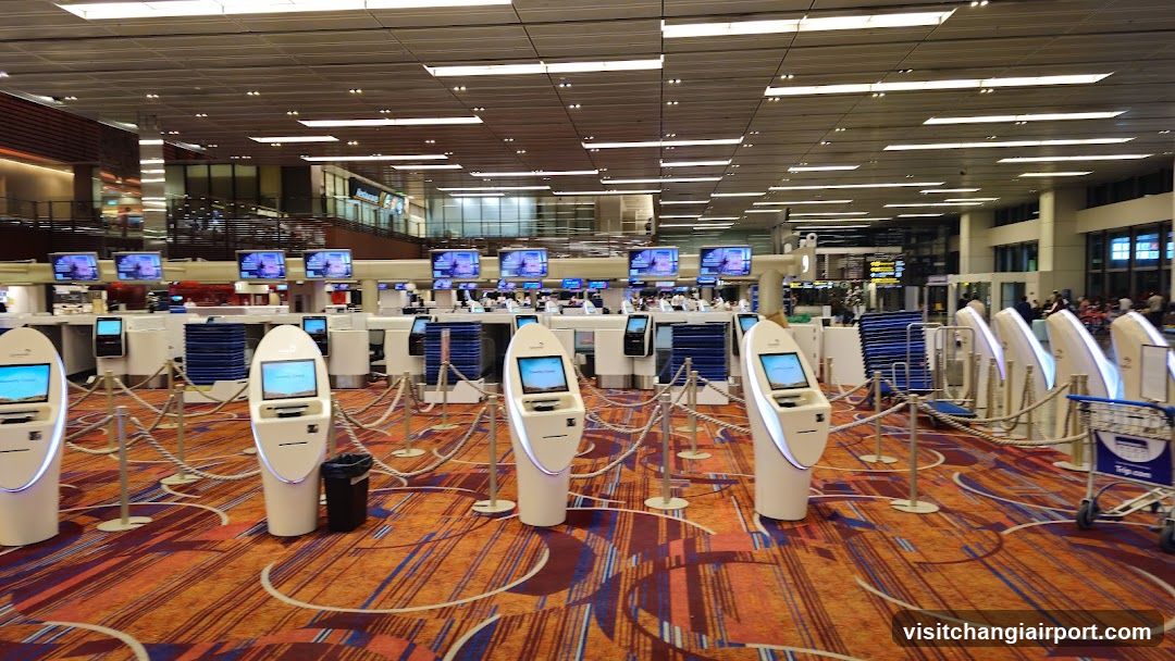 The image shows a spacious airport terminal check-in area featuring multiple self-service kiosks arranged in rows, each equipped with touchscreens and illuminated bases. The floor is covered with patterned carpeting in warm tones of orange, red, and blue. Stanchions guide passengers toward the counters in the background, where staff members are visible behind desks displaying monitors. Overhead lighting illuminates the high ceiling with recessed panels. A few luggage carts are parked near the right side. No signage or menus are present; this is an interior operational space.