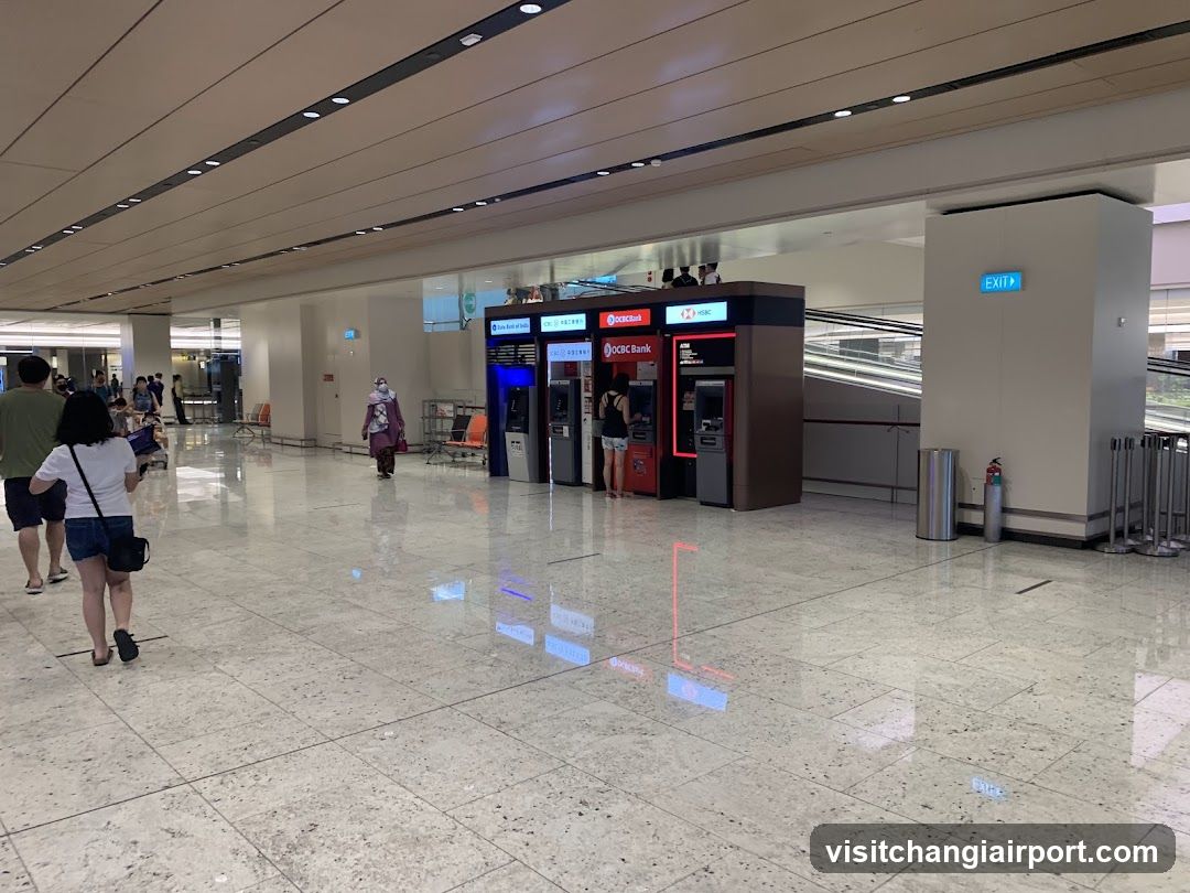 A spacious, well-lit indoor area with polished marble flooring and recessed ceiling lighting. In the center, there is a row of ATMs from OCBC Bank and other banks, including one with red branding. A person in a purple uniform walks nearby, while others are seen in the background near seating areas. An exit sign is visible on the right wall.