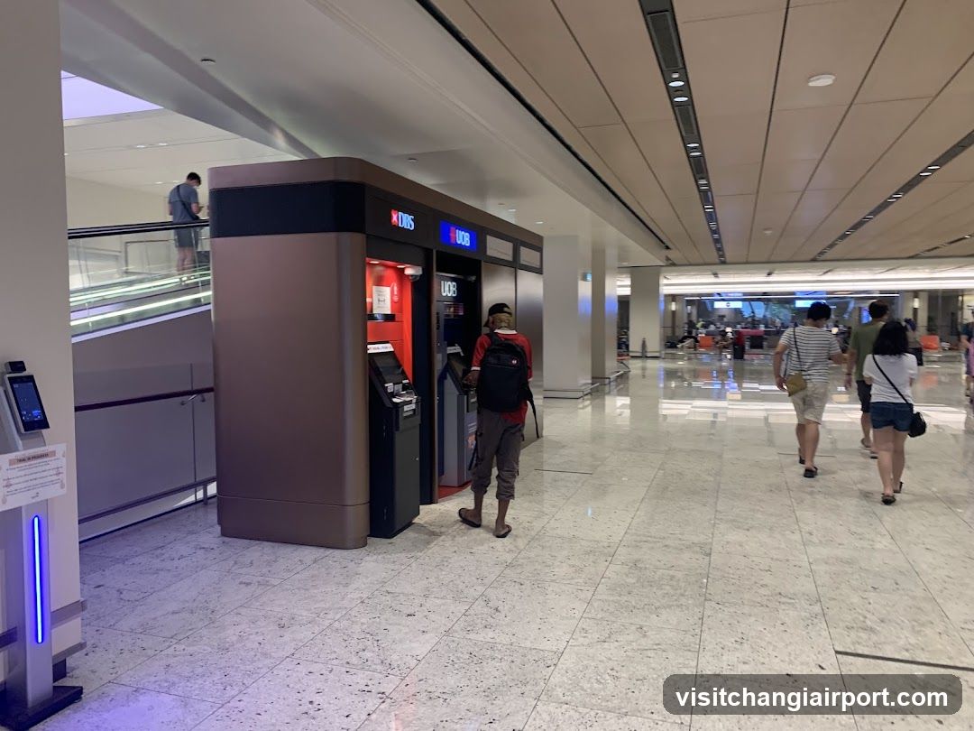 A spacious airport terminal hallway with polished marble flooring and recessed ceiling lighting. A person wearing a red shirt and backpack stands near an ATM kiosk labeled 'UBS'. In the background, travelers walk toward the terminal exit or gate area. An escalator is visible on the left side.