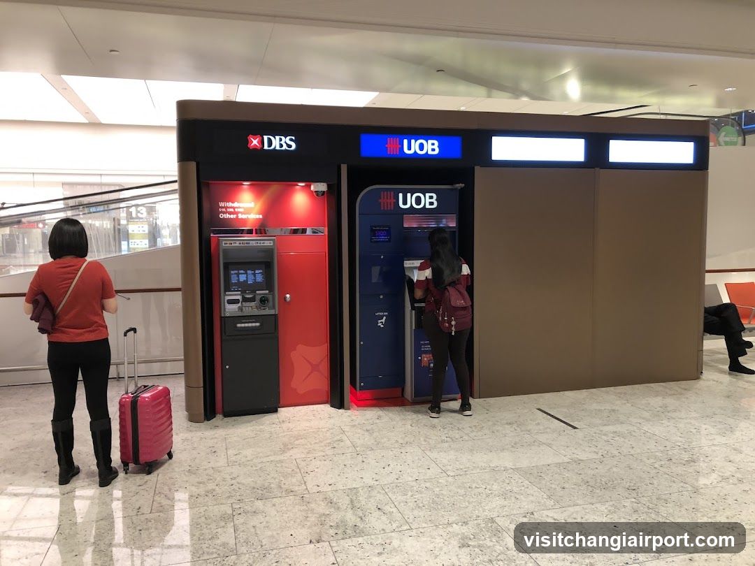 A modern ATM kiosk with DBS and UOB branding, featuring two ATMs under a covered structure. A woman in red stands near the DBS ATM with a pink suitcase, while another person uses the UOB ATM. The setting appears to be an airport terminal with polished floors and visible signage.
