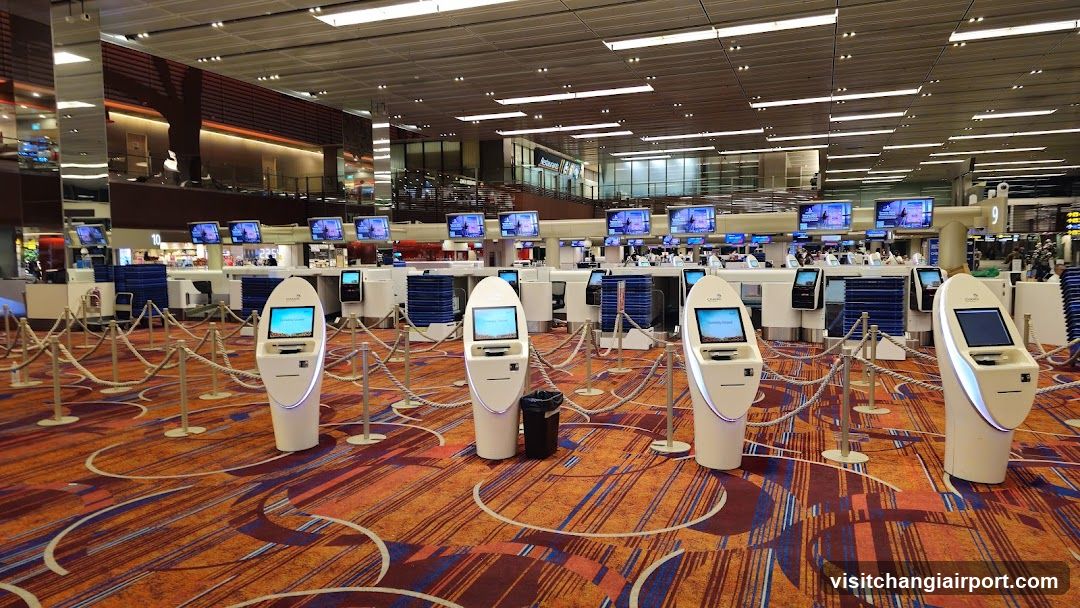 A spacious airport terminal interior featuring multiple self-service kiosk stations arranged in rows on a patterned carpet with orange and blue tones. The kiosks have illuminated screens and are separated by stanchions and ropes. In the background, there is an elevated walkway with glass railings, additional service counters, and digital display boards showing flight information or advertisements. Lighting is bright and modern, with recessed ceiling lights and long fluorescent fixtures.
