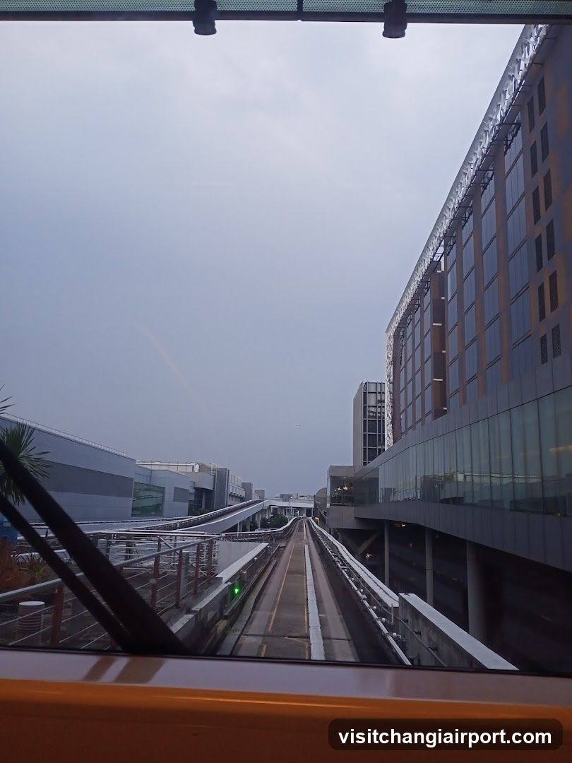 View from inside a monorail looking forward along elevated tracks toward airport terminal buildings under an overcast sky.