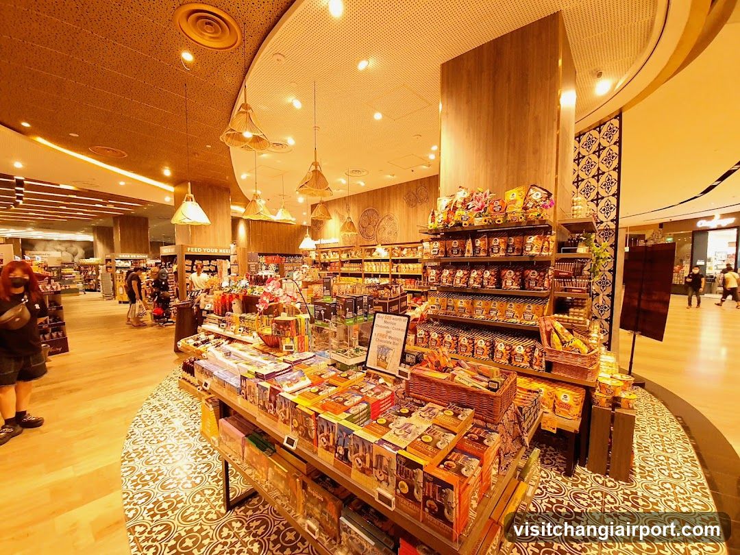 Interior of a brightly lit retail store with wooden accents and hanging pendant lights, featuring shelves stocked with packaged goods, snacks, and merchandise displayed on tiered stands.