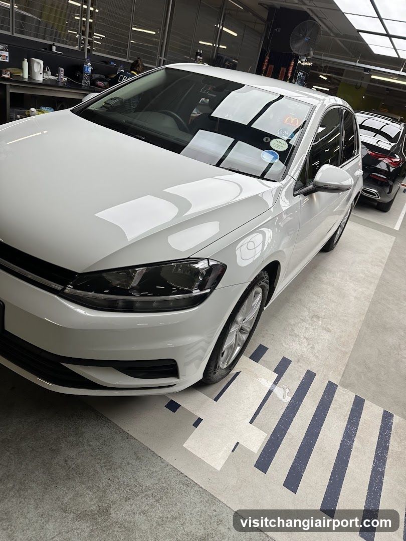 A white car is parked inside a workshop or garage, with tools and equipment visible in the background.