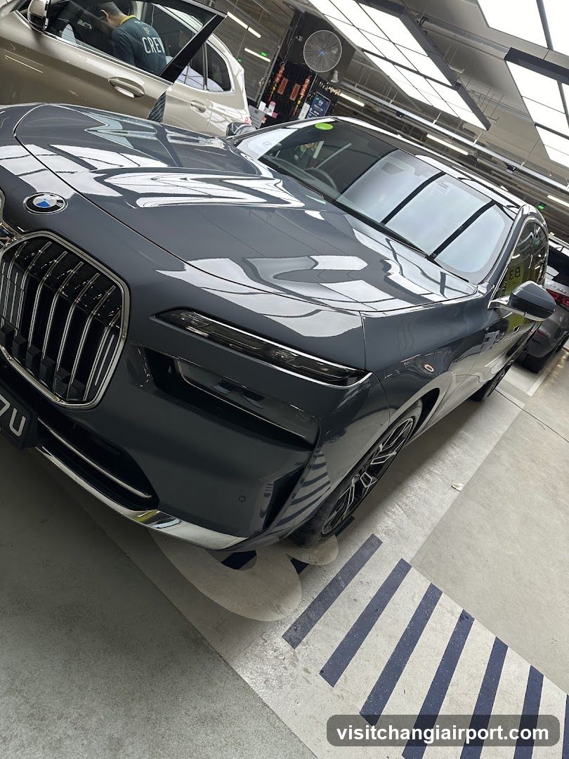 A close-up view of a sleek, dark gray BMW vehicle parked inside a well-lit garage or showroom. The car's glossy exterior reflects the overhead lights and surrounding environment. In the background, another vehicle is partially visible with its door open, suggesting an active service or display area. The setting appears to be a professional automotive facility.