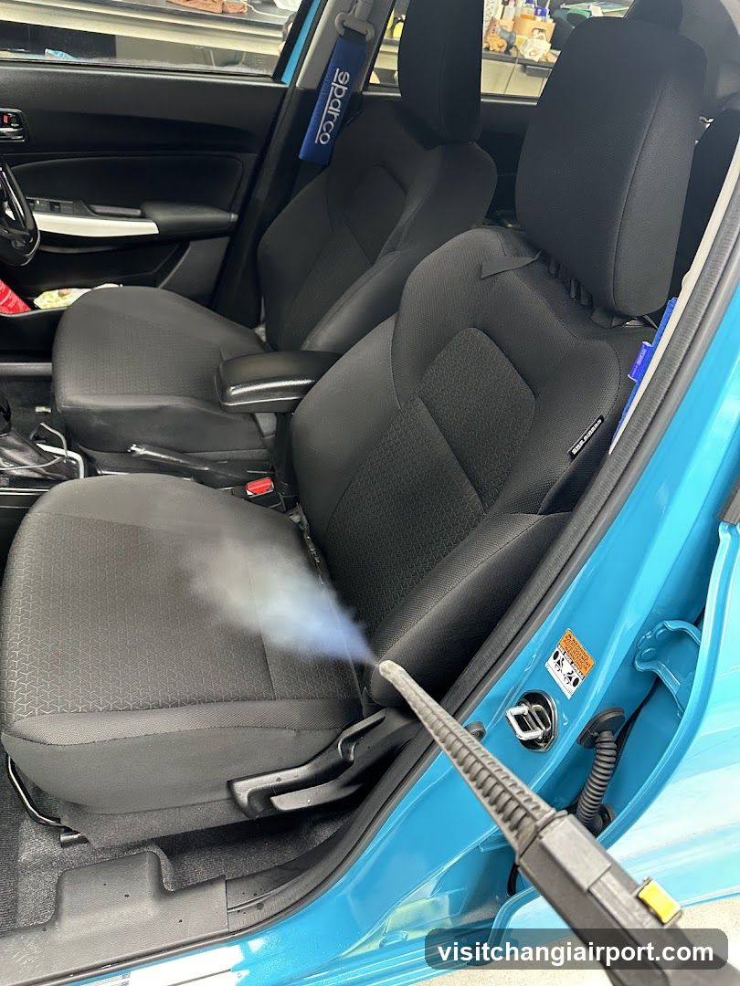 A close-up view of a car's interior, focusing on the driver and passenger seats being cleaned with a steam cleaner. The car has black fabric seats with a textured pattern, and blue Sparco seatbelt covers are visible. A handheld steam cleaning device is spraying steam onto the passenger seat area. The car door is open, revealing part of the exterior paint which appears to be light blue or turquoise. Various items like bottles and containers can be seen in the background through the window.