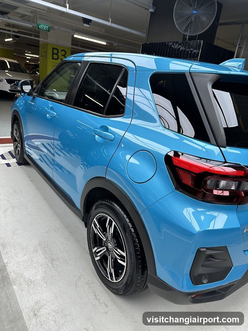 A bright blue compact SUV is parked inside a well-lit underground parking garage. The vehicle features black trim around the wheel arches and lower body, with multi-spoke alloy wheels. In the background, another white car is partially visible near an exit sign labeled 'EXIT' and a yellow pillar marked 'B3'. A large industrial fan hangs from the ceiling.