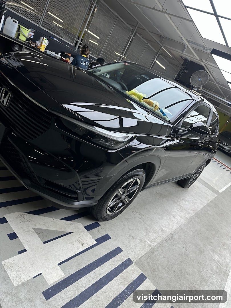 A black Honda HR-V is parked inside a detailing or car wash facility. The vehicle's glossy finish reflects the overhead lights and surroundings. A yellow microfiber cloth rests on the windshield, suggesting recent or ongoing cleaning. In the background, another person wearing a 'CREW' shirt works near a counter with various bottles of detailing products. The floor features blue and white striped markings, typical of automotive service bays.