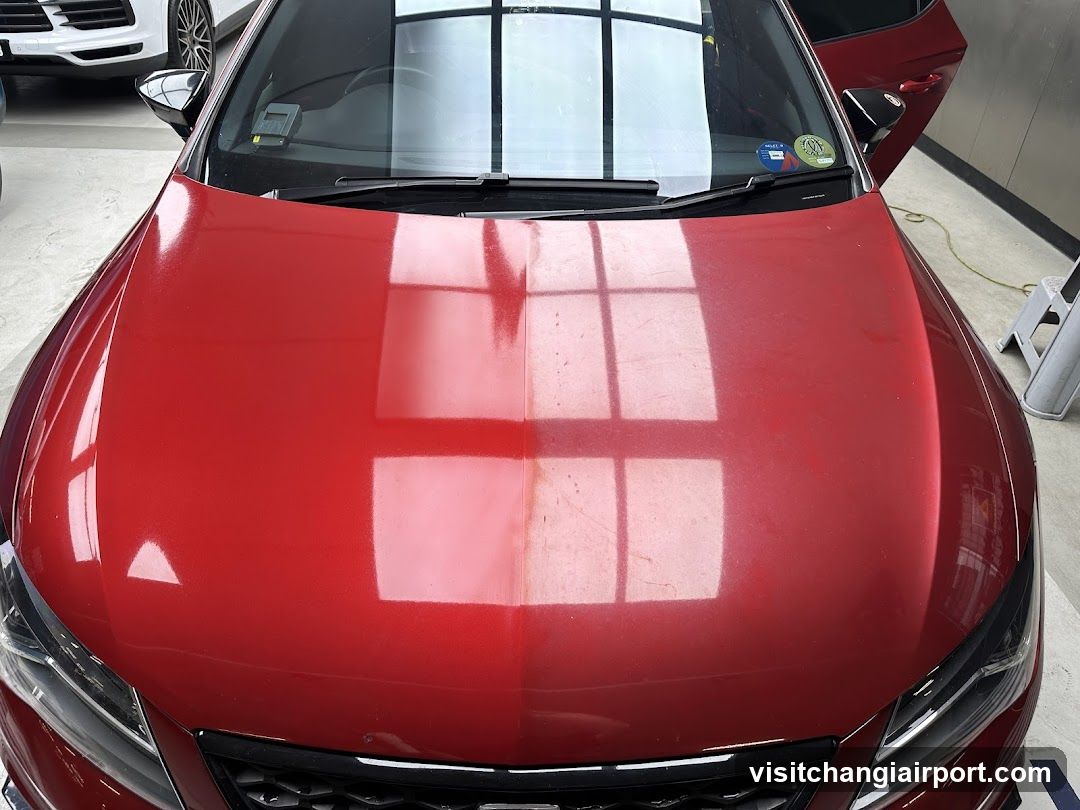 A close-up view of a red car's hood, showcasing reflections from overhead lights and windows in what appears to be an indoor showroom or garage setting.