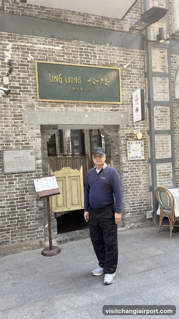 A man stands in front of a brick building with a sign reading 'LING LOONG | 伶隆' and the year '1923'. The entrance features an arched doorway with a wooden door partially open. A menu board is placed to the left of the entrance, displaying some text.