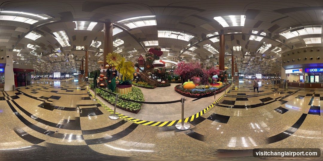 A wide-angle view of a spacious airport terminal hall featuring polished terrazzo flooring and high ceilings with recessed lighting. In the center, there is an elaborate seasonal display resembling a whimsical garden or fairy tale scene, complete with oversized flowers, colorful structures, and stanchions cordoning off the area. On the right side, a shop front displays signage reading 'U08' above its entrance.