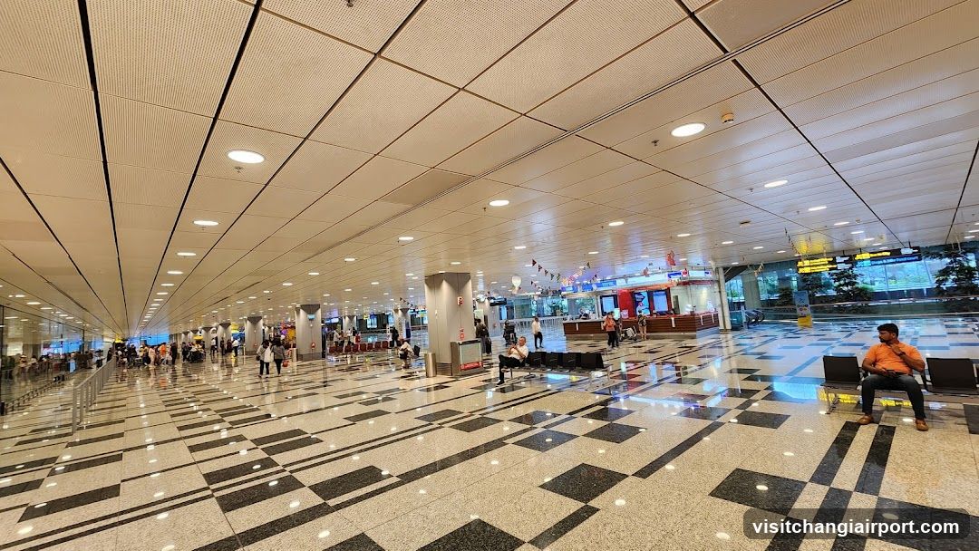 A spacious airport terminal with a checkered floor, rows of seating, and passengers waiting. In the background, there are service counters and digital displays showing flight information. A person in an orange shirt sits on the right side.