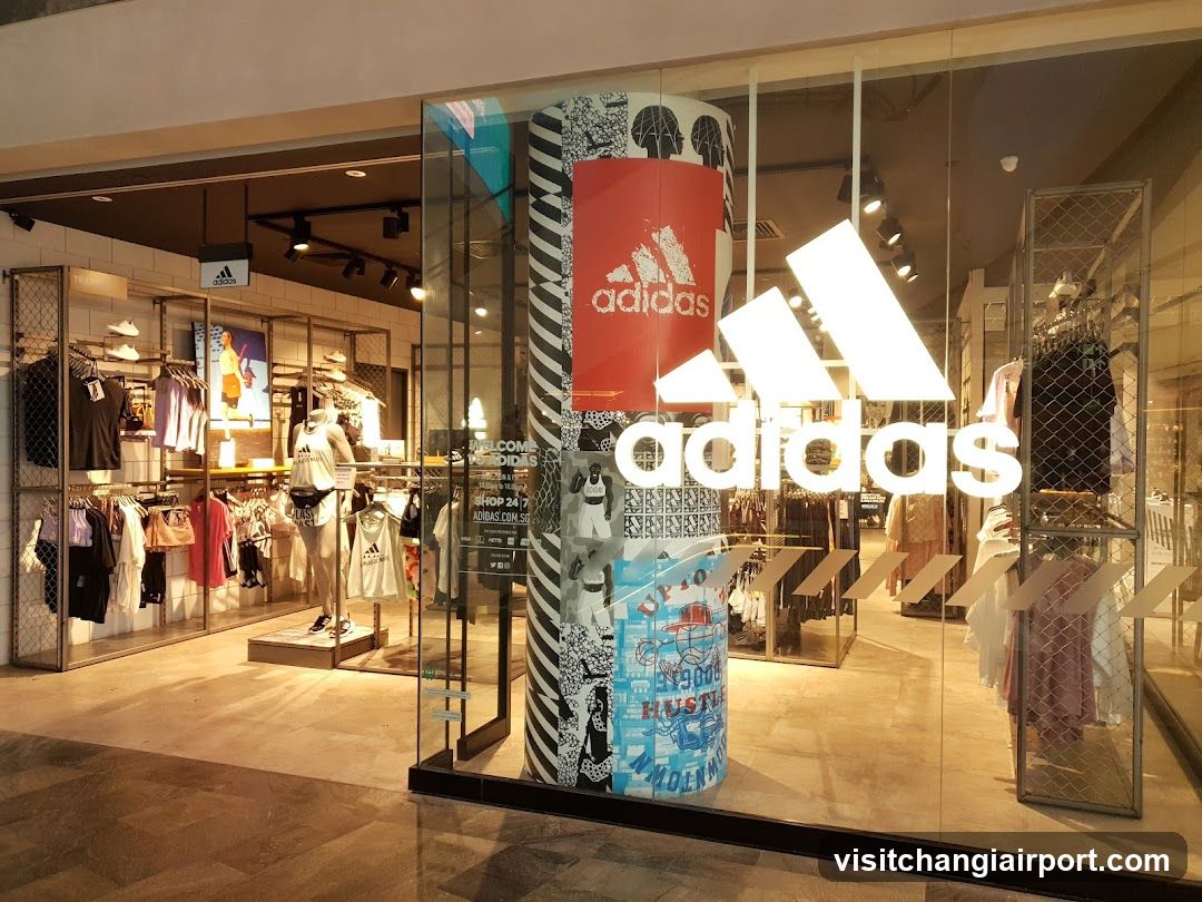 The image shows the exterior of an Adidas store with a large illuminated logo on the glass facade. Inside, clothing racks display various apparel items including shirts and dresses. A cylindrical pillar features colorful graphics and branding elements.