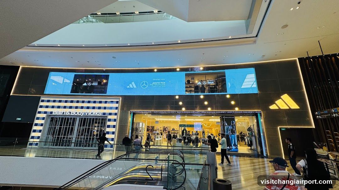 The image shows the exterior of several shop fronts in a modern shopping mall. Visible brands include Bath & Body Works, Mercedes-Benz, and Adidas. The storefronts feature illuminated signage and glass entrances with people walking by.