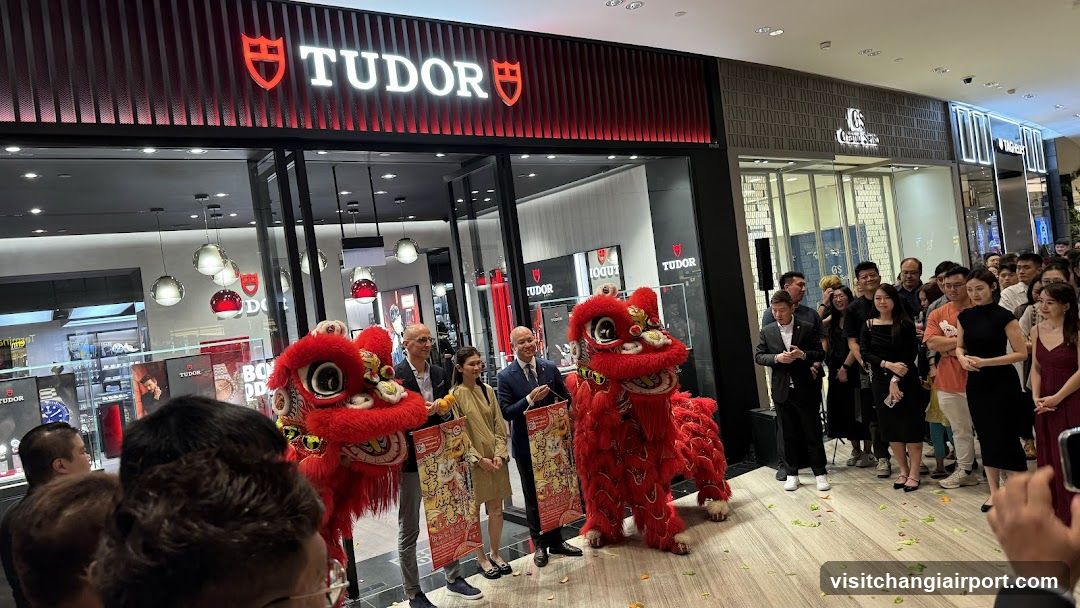 A Tudor watch store storefront with red and black signage featuring the Tudor logo. Two red lion dance performers are present in front of the entrance, surrounded by a crowd of people celebrating the opening. Confetti is scattered on the floor.