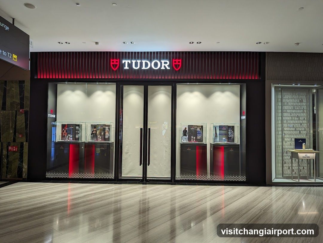 A modern shop front with a black facade and red vertical slats above the entrance. The Tudor logo is prominently displayed in white letters flanked by two red shield emblems. Large glass windows showcase watches on display stands, illuminated from within. The floor is polished wood, reflecting the store's interior lighting.