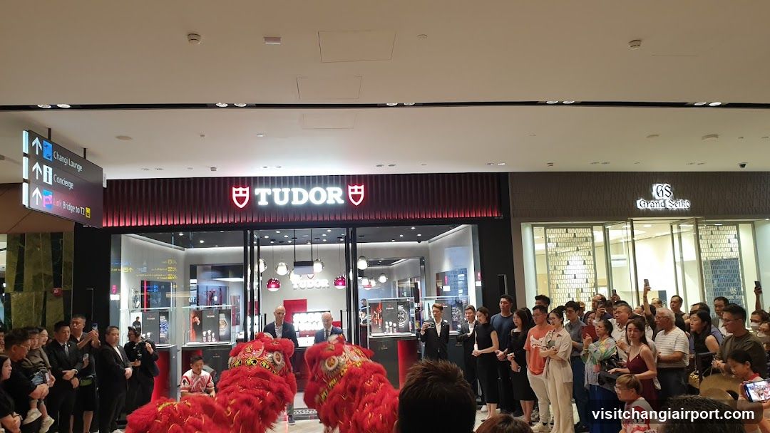 A storefront with a red and black facade featuring the 'TUDOR' logo illuminated in white letters above glass doors. A crowd of people is gathered outside, some holding phones to take photos or videos. Two red lion dance costumes are visible near the entrance. Signage on the left points to Chang Lounge, Concourse, and Exit Bridge to T2.