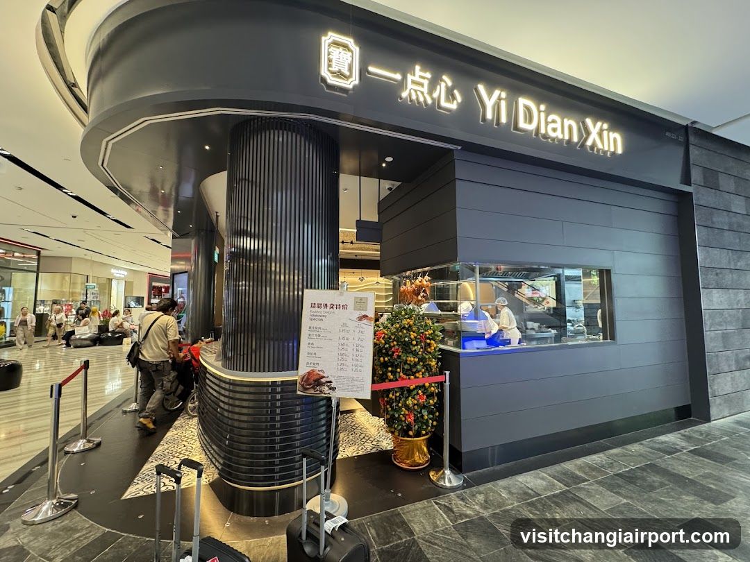 Modern dark gray shop with illuminated signage reading Yi Dian Xin and Chinese characters. Large glass window shows kitchen staff in white uniforms preparing food. Decorative potted plant near entrance. Stanchions guide customer flow. Luggage visible in foreground suggesting travel or airport setting.