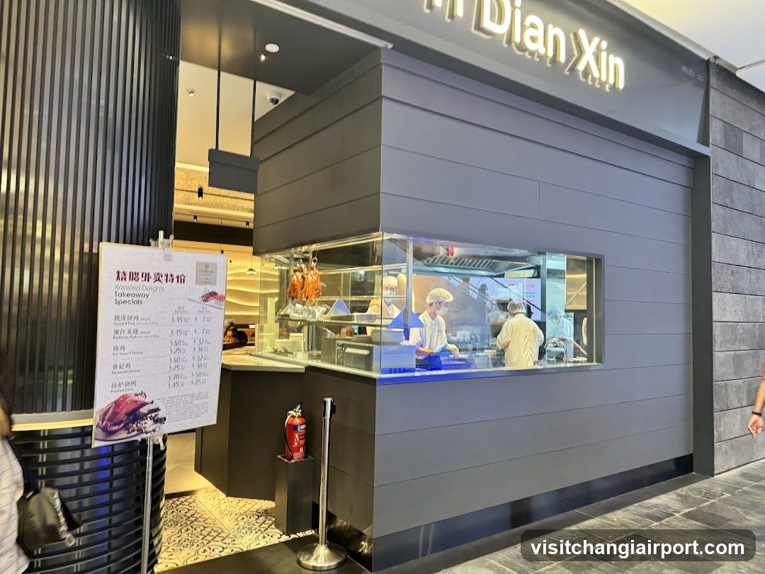A modern shop front with a gray facade and illuminated signage reading 'Dian Xin'. Inside, staff in white uniforms are preparing food behind a glass counter. A menu board is visible on the left side of the entrance.