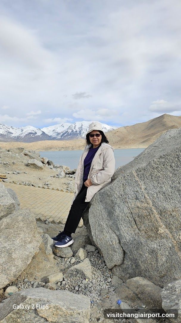 Person at a rocky lakeside with snow-capped mountains in the background