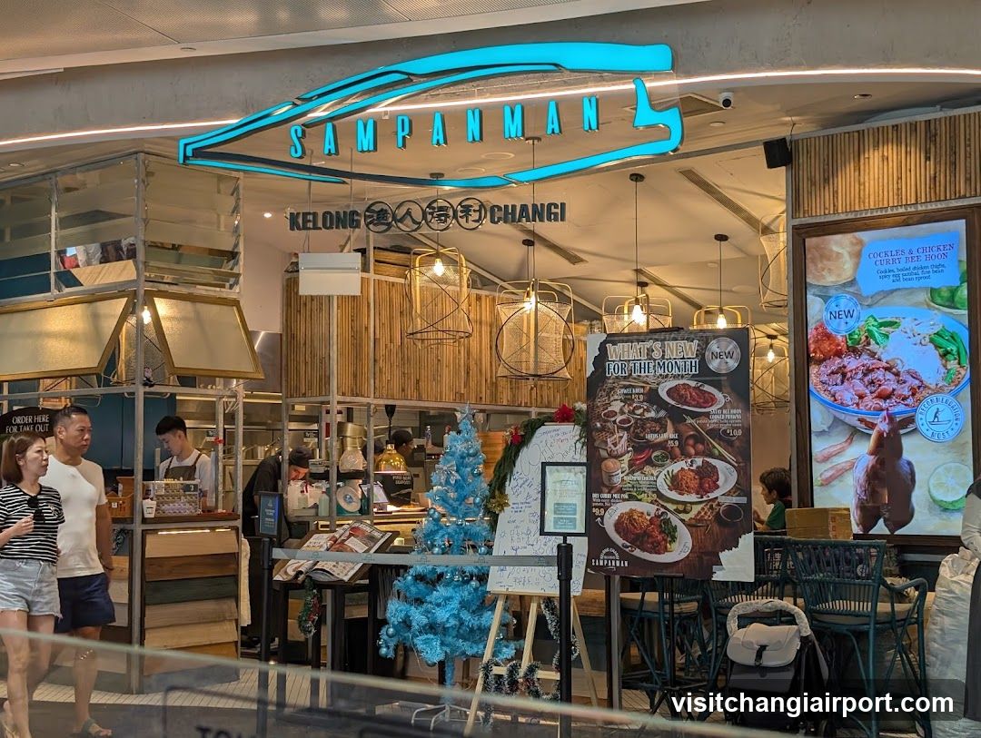 A brightly lit storefront with a neon blue sign reading 'SAMPANMAN' and 'KELONG 深人海味 CHANGI'. The entrance features wooden paneling, hanging pendant lights, and a small blue Christmas tree. Staff are visible behind the counter, and customers stand near the entrance. A large poster advertises new menu items including 'Cockles & Chicken Curry Hot Pot', while another board displays various dishes.
